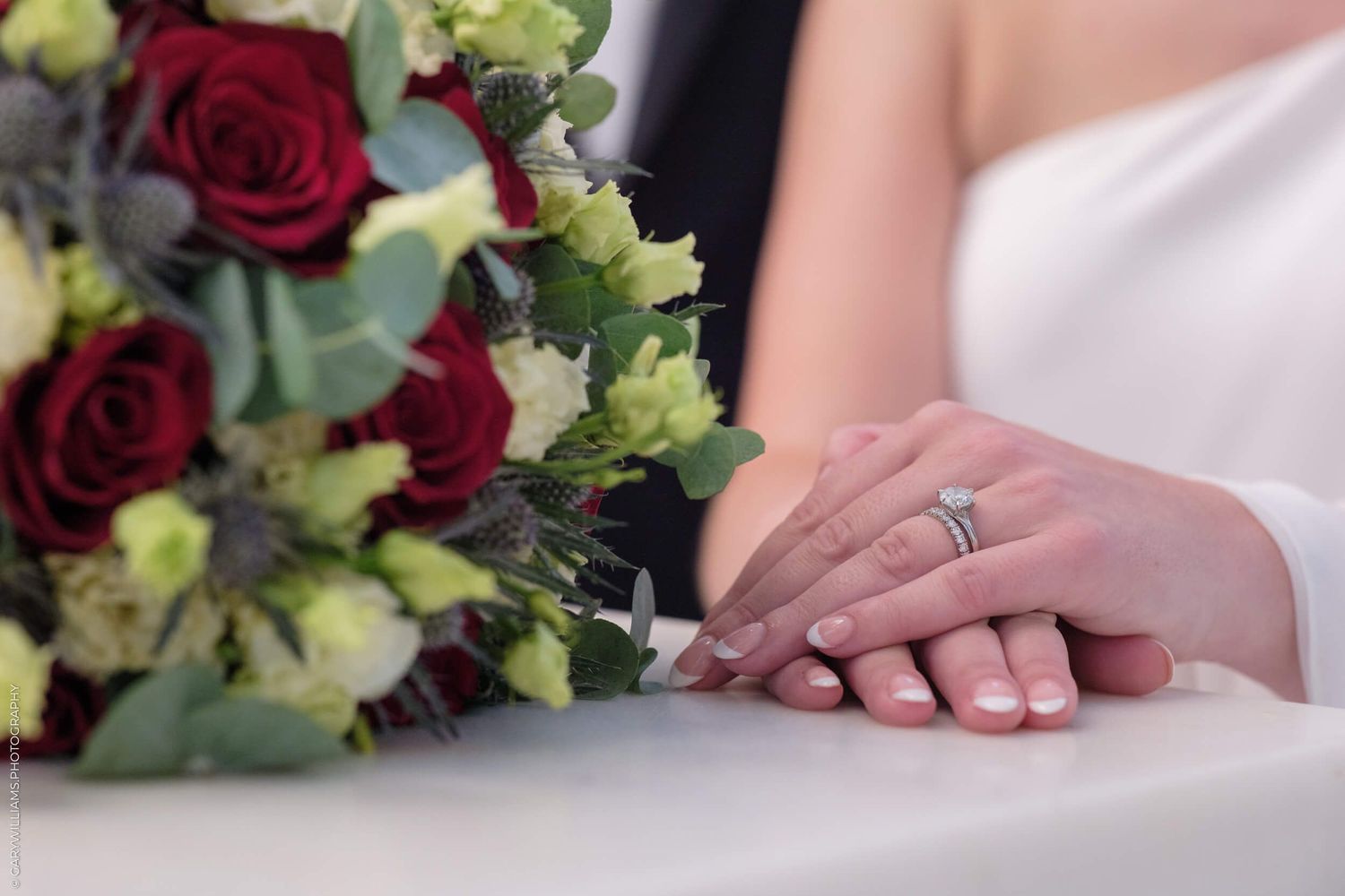 Wedding detail showing clasped hands wearing rings next to a red rose and greenery bouquet.