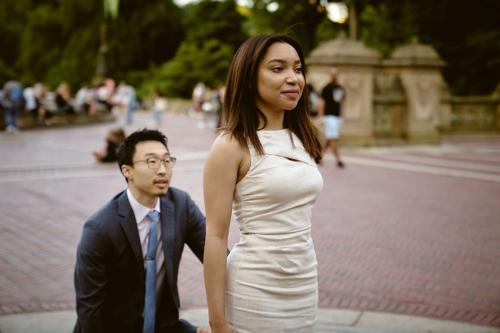 A couple walking together on a brick plaza, with the woman wearing a white dress and the man in a navy suit. surprise engagement