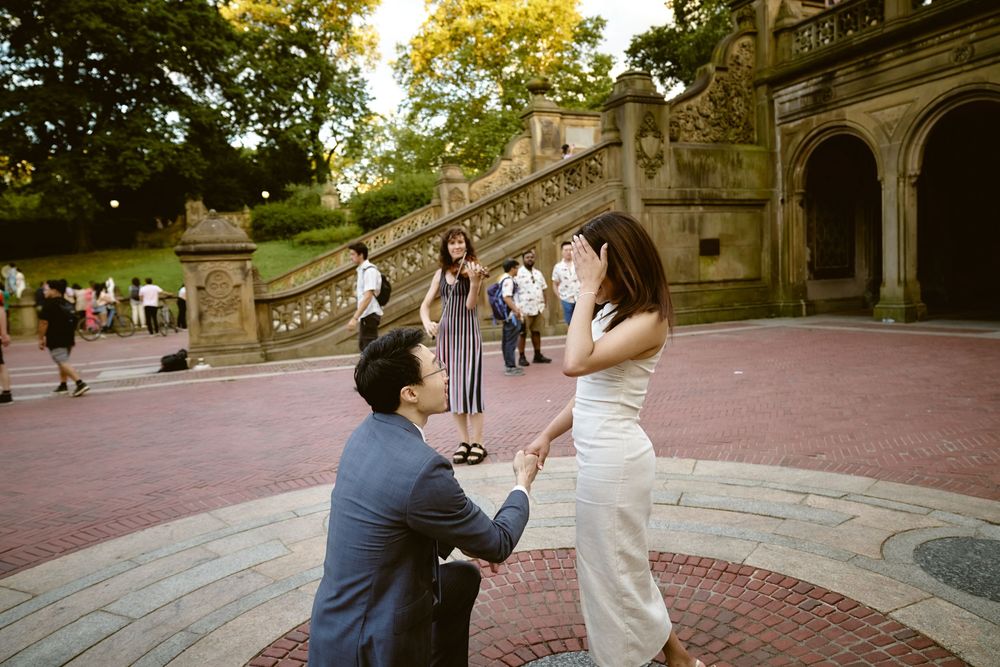 A romantic proposal moment taking place on the historic Bethesda Terrace steps in Central Park during golden hour. Surprise engagement in ce