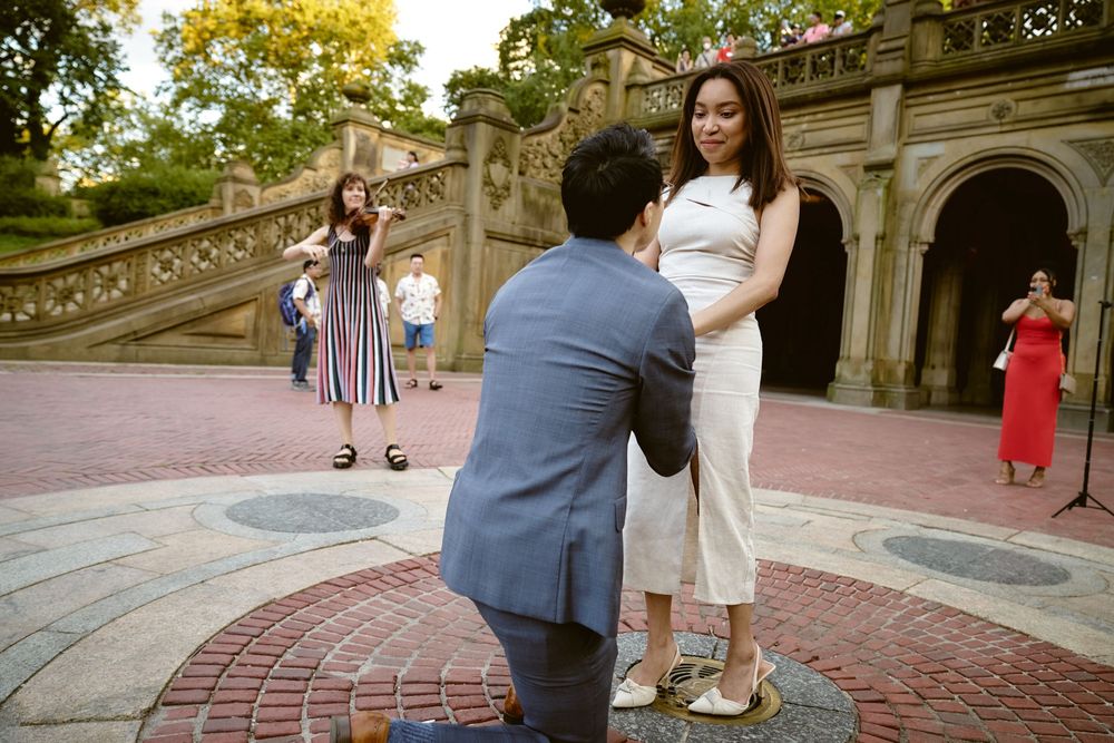A romantic proposal scene taking place on an ornate stone terrace near historic archways in Central Park. Surprise engagement in central par