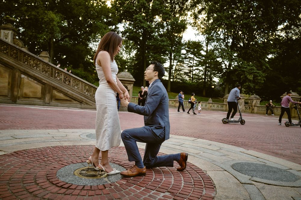 Wide panorama of a romantic proposal in a park at sunset with figures kneeling on a paved circular area. Surprise engagement in central park