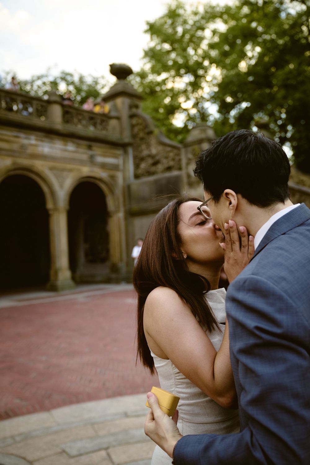 Romantic kiss between couple in front of stone archway during outdoor engagement session at dusk. Surprise engagement in central park