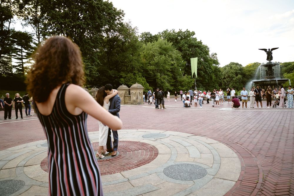 A person in a striped dress looks towards Bethesda Fountain in Central Park on a busy summer day. Surprise engagement in central park
