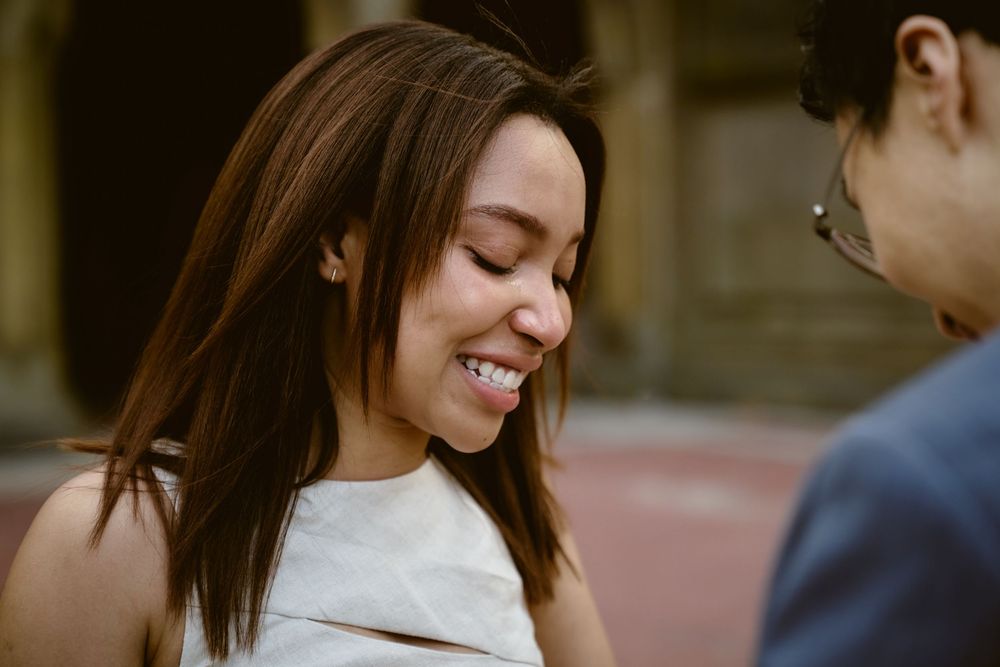 Two people share a candid moment outdoors, with one wearing a white top and smiling joyfully. Surprise engagement in central park
