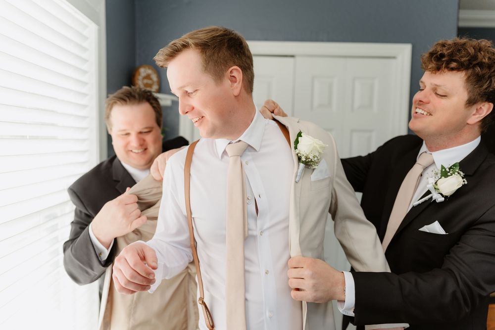 Groomsmen help with wedding preparations in a light-filled room while sharing a candid moment together.