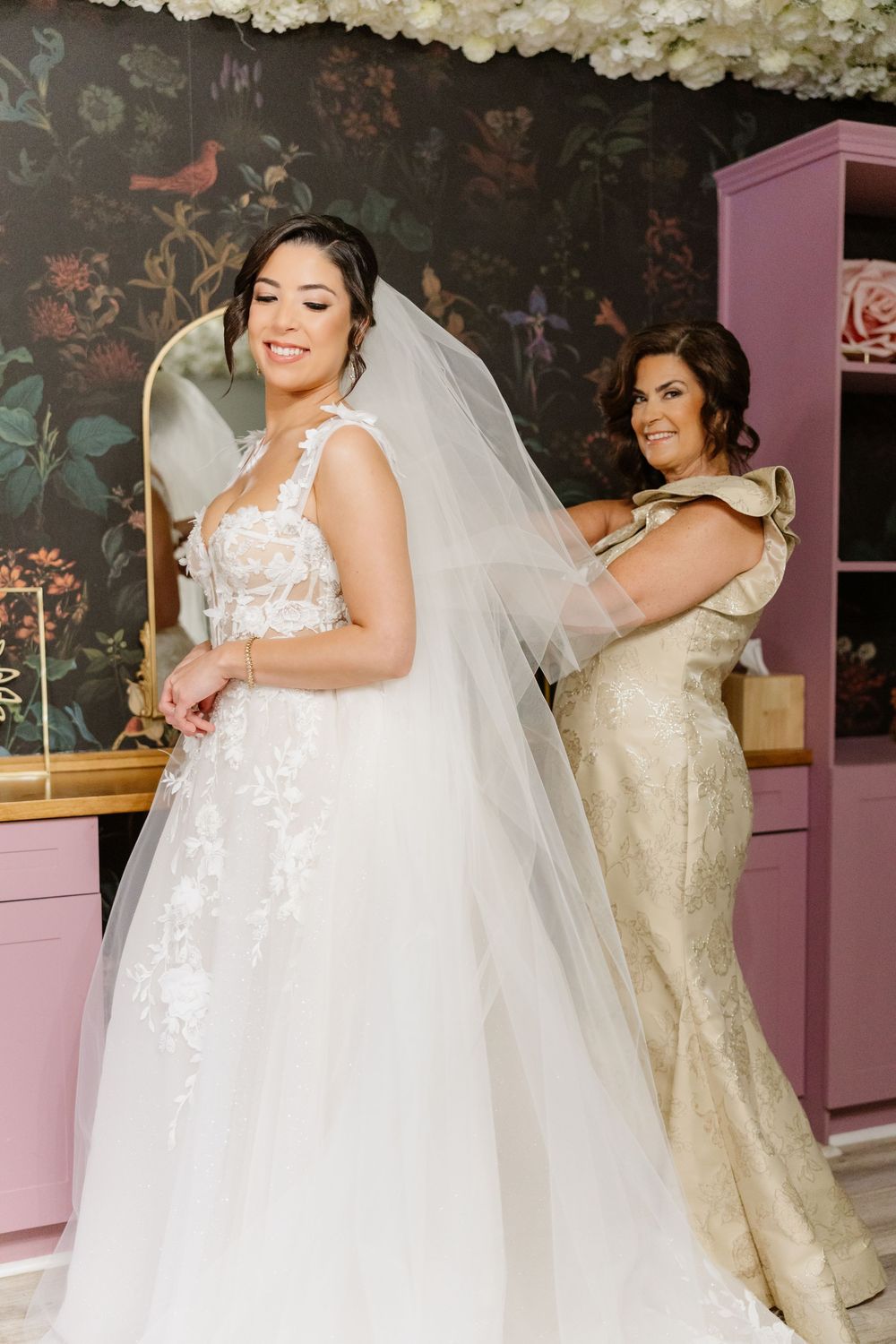 A wedding dress fitting moment captured against floral wallpaper with gold accents.