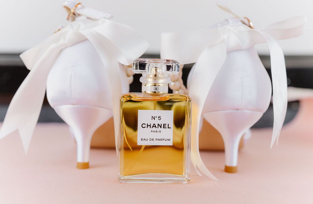 Wedding accessories including white heels and Chanel perfume bottle arranged on pink surface.