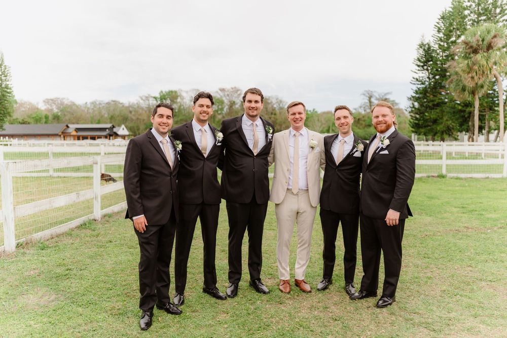 A groom in a light suit stands with five groomsmen in dark suits on a rustic farm property with white fencing.