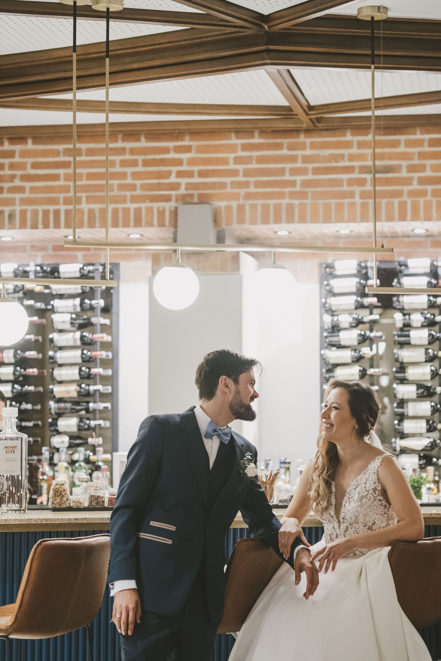 Bride and groom share joyful laughter while sitting together in a restaurant with modern wine racks in background.