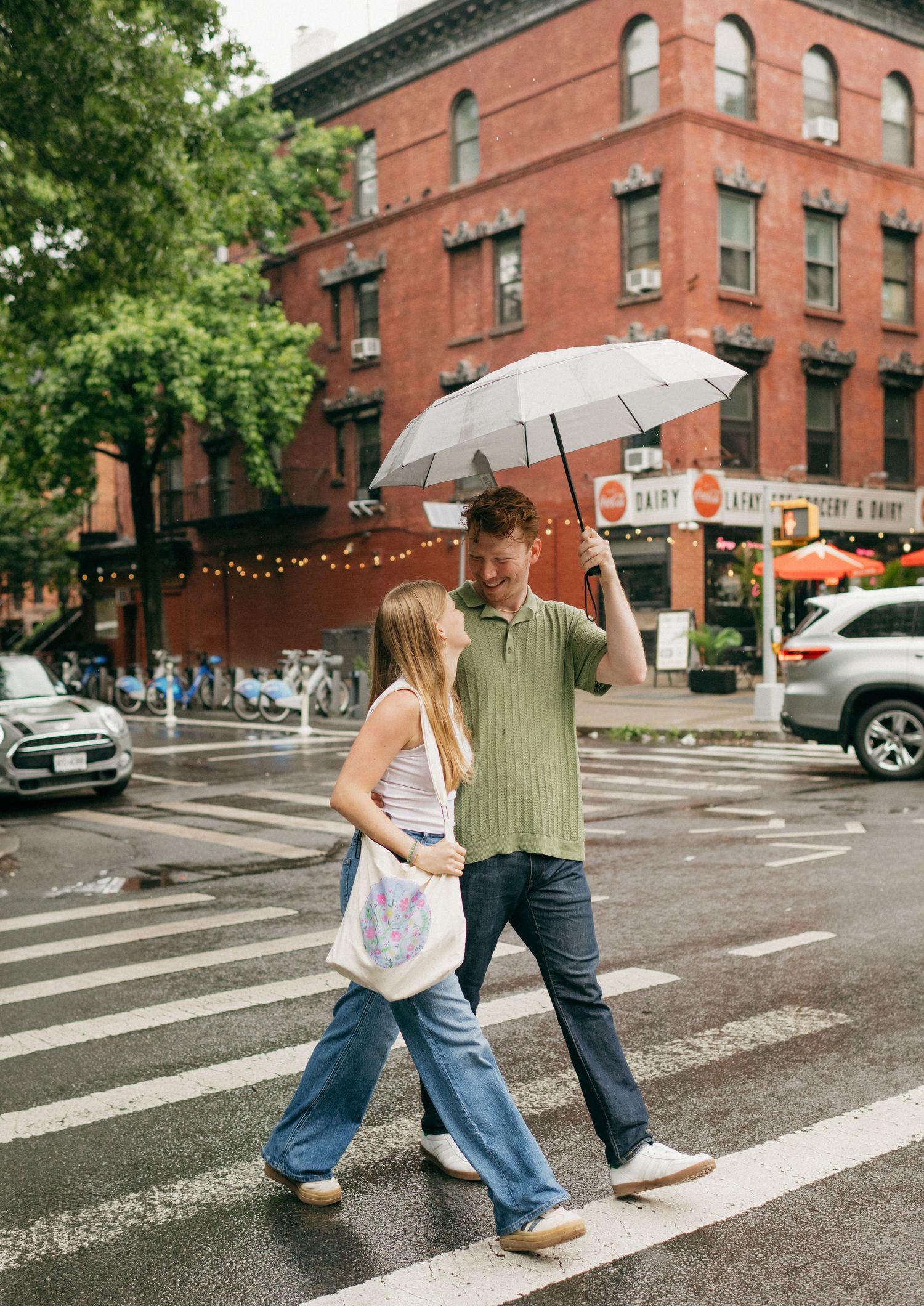 Couple crosses street with umbrella in front of historic brick buildings on a rainy day.