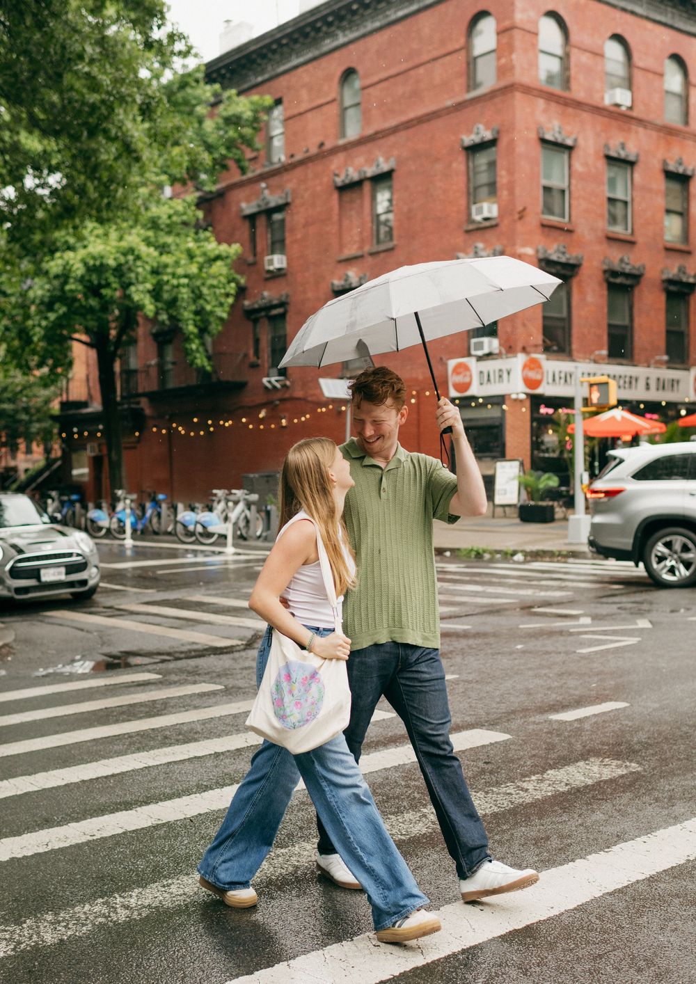 Couple crosses street with umbrella in front of historic brick buildings on a rainy day.