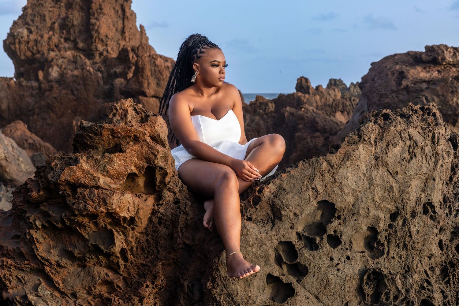 Nayesha poses for a portrait session on rocky terrain, wearing a white dress, with a serene expression against a beautiful sky backdrop.