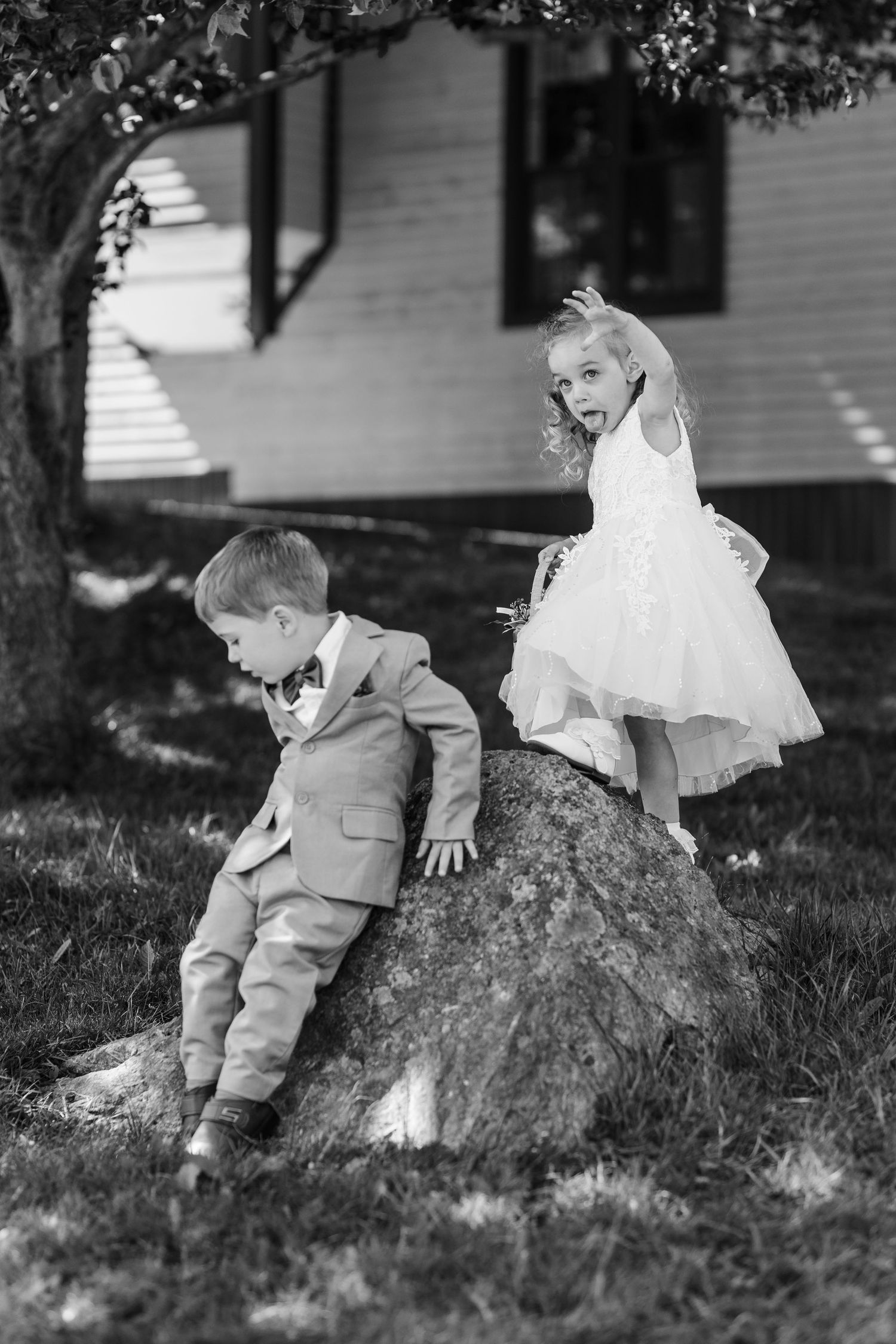 Black and white photo of children playing on a large rock in formal attire, dress and suit.