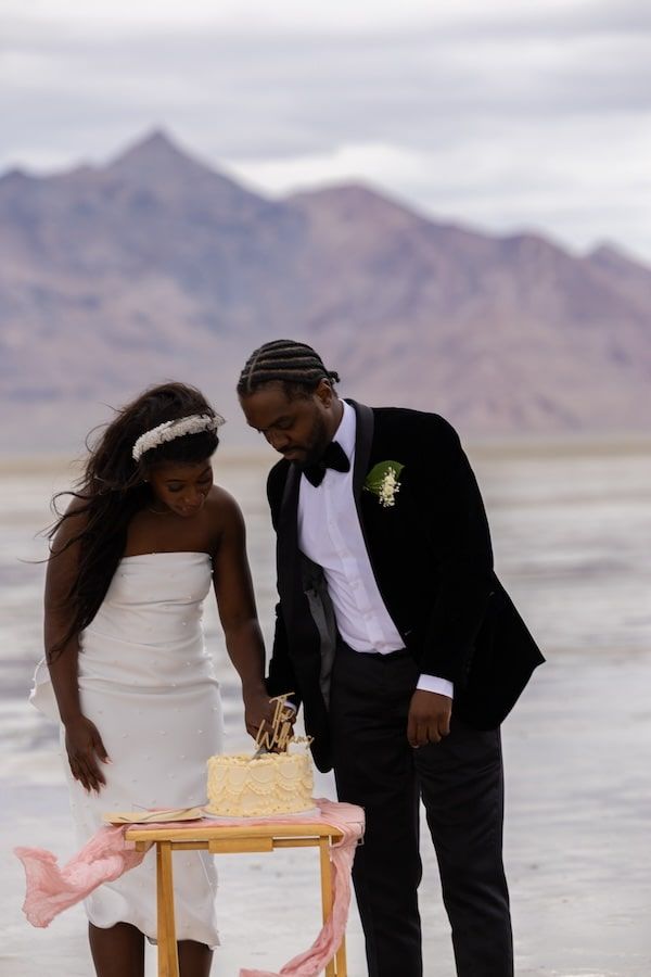 A couple leans forward over a small wooden table on a desert salt flat with mountains in the background.