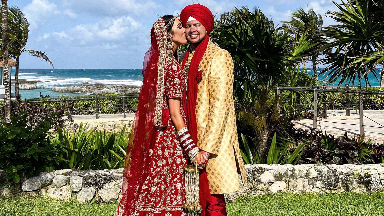 A couple in traditional Indian wedding attire poses together on a scenic beach with palm trees and ocean views.