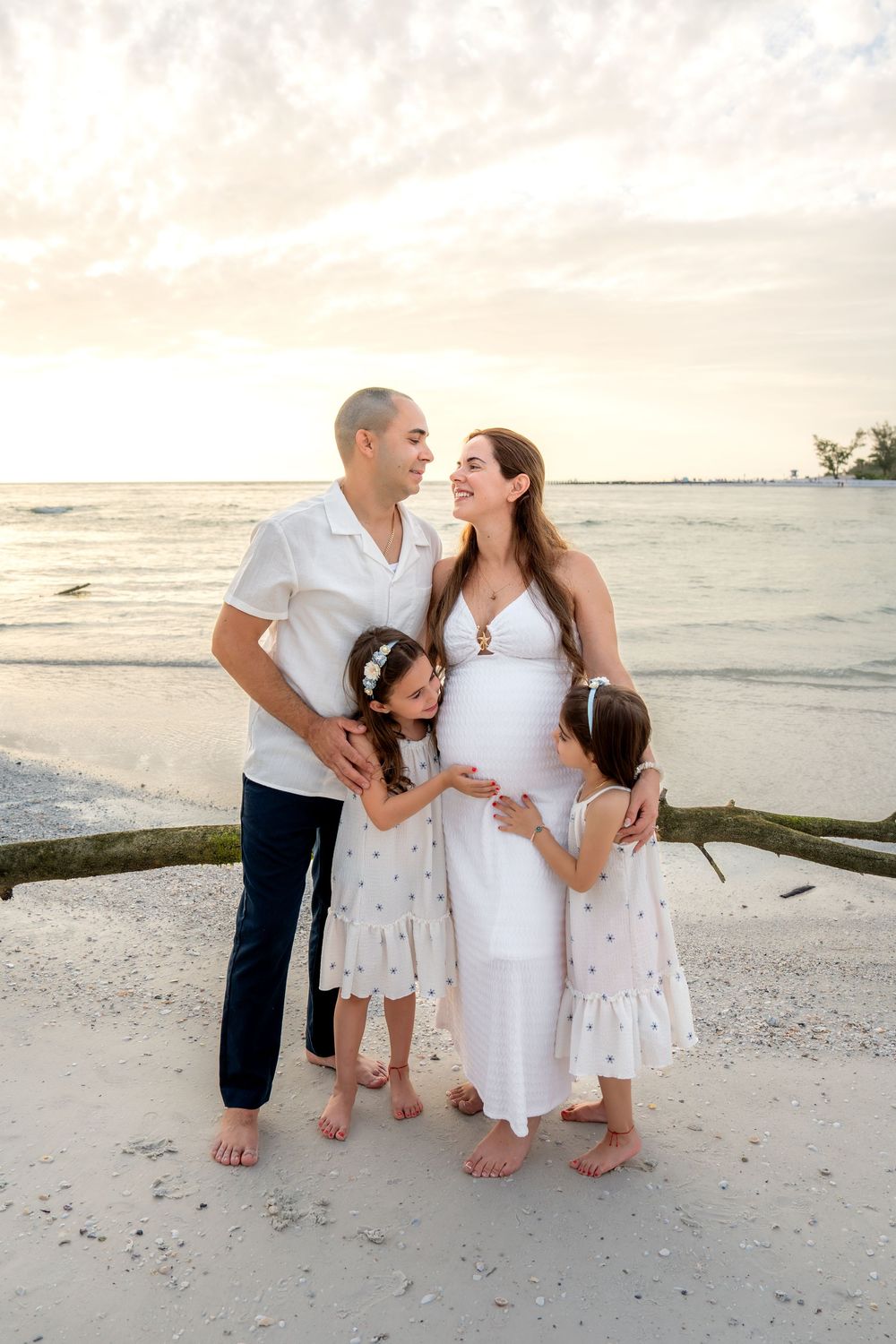 Family portrait on the beach at sunset, pregnant mother in white dress with husband and two young daughters.
