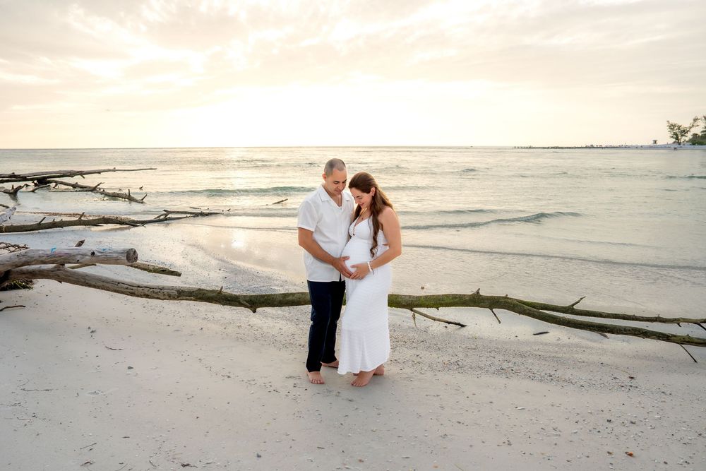 Expecting couple standing together on a sandy beach at sunset, surrounded by driftwood and calm ocean waters.