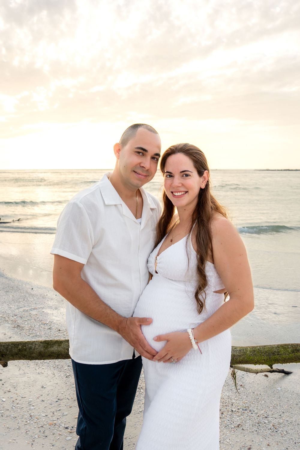 Smiling pregnant couple posing on the beach at sunset, both cradling the baby bump in white outfits.