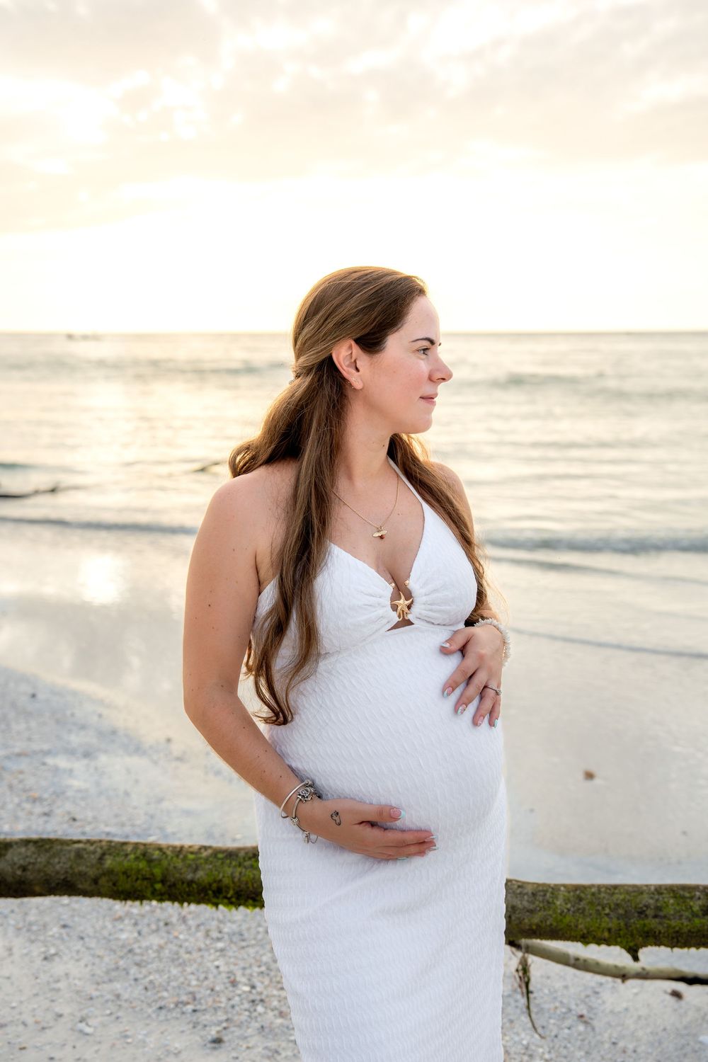 Pregnant woman in white dress cradling her baby bump on the beach at golden hour, gazing peacefully.