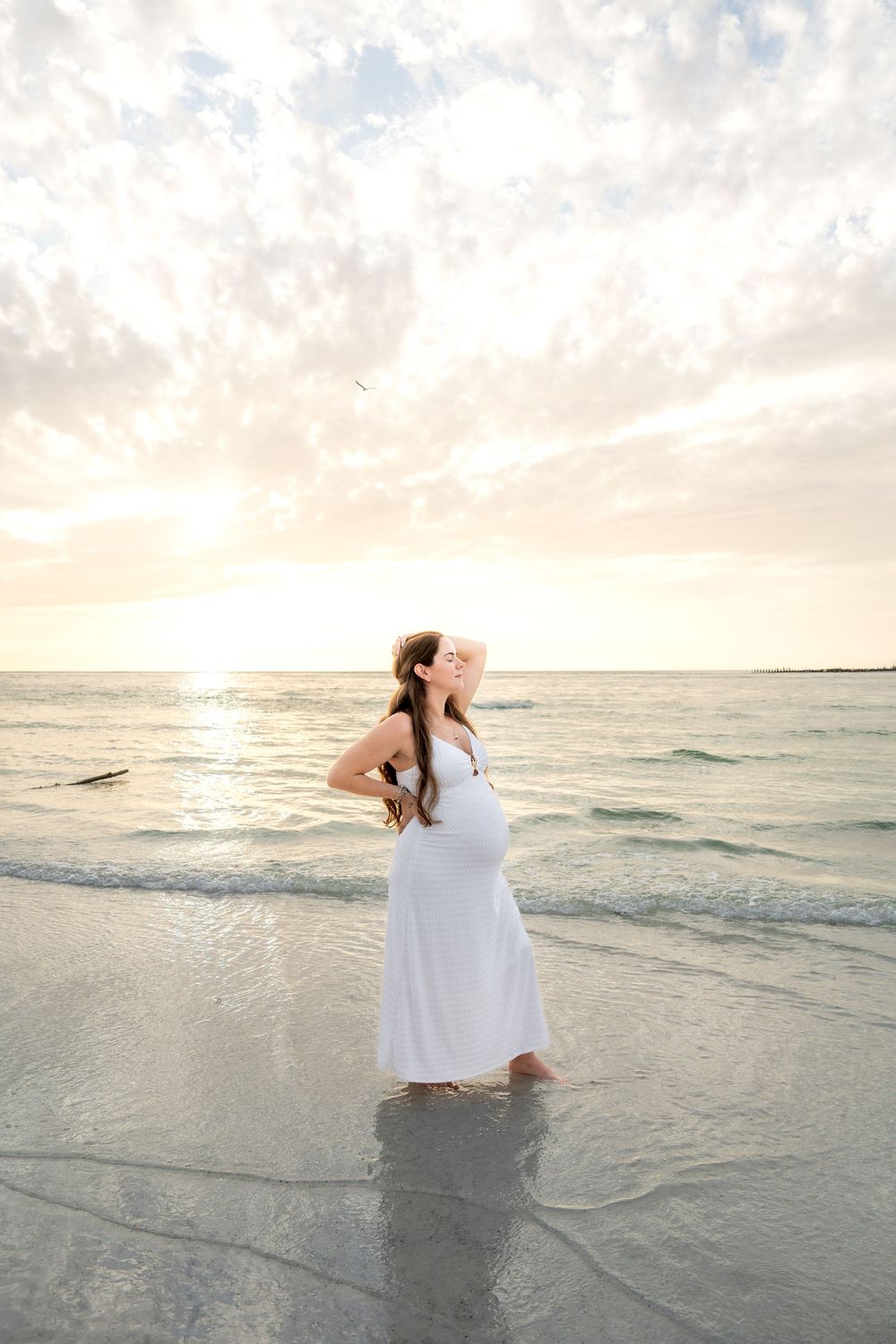 Pregnant woman in white dress standing in shallow ocean waves at sunset, looking up at a cloudy sky.