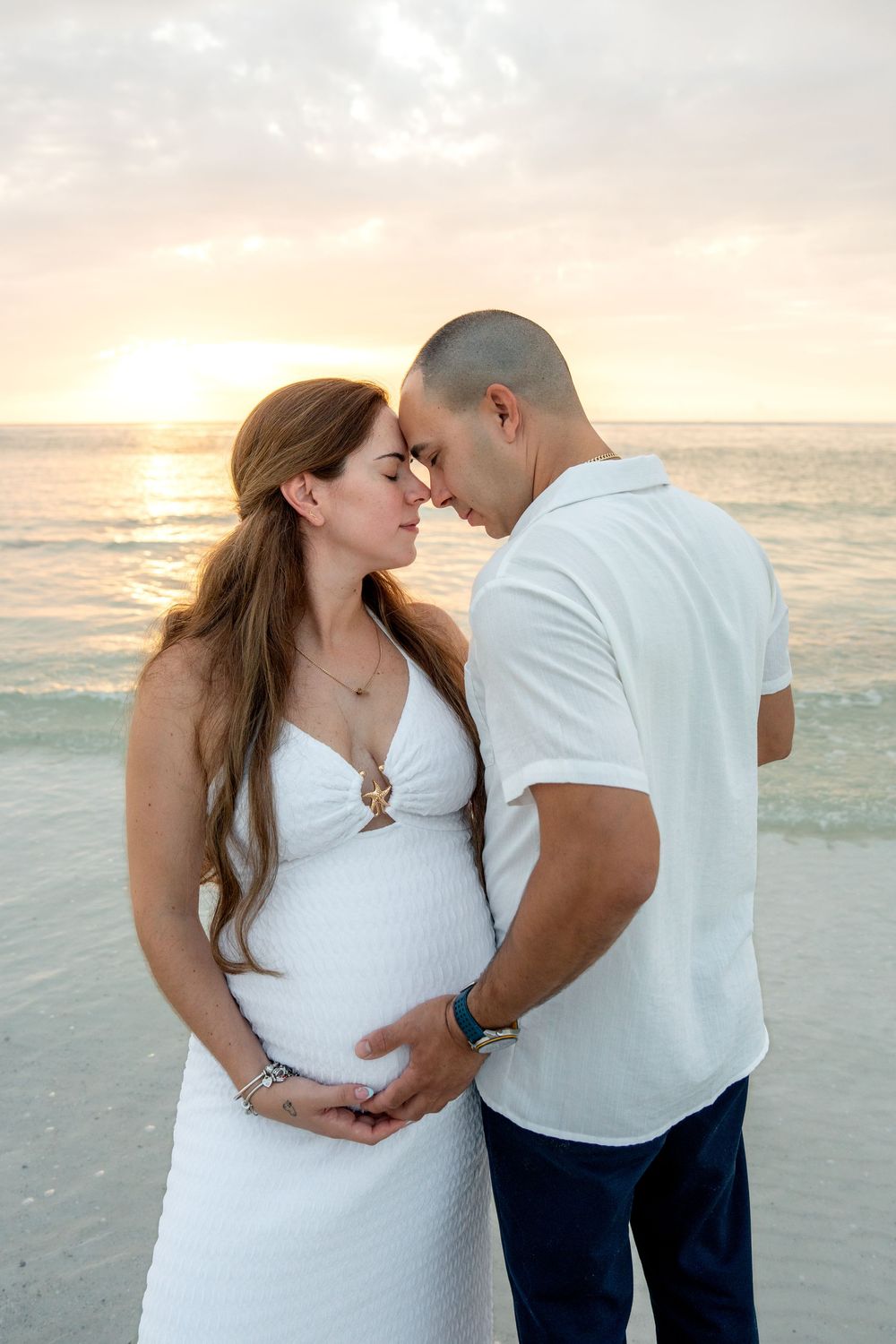 Pregnant couple in white touching foreheads tenderly on a beach at sunset.
