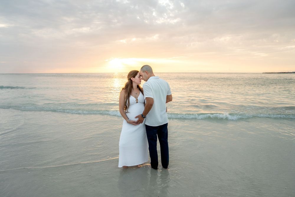Expecting couple standing in shallow ocean water holding baby bump at golden sunset.