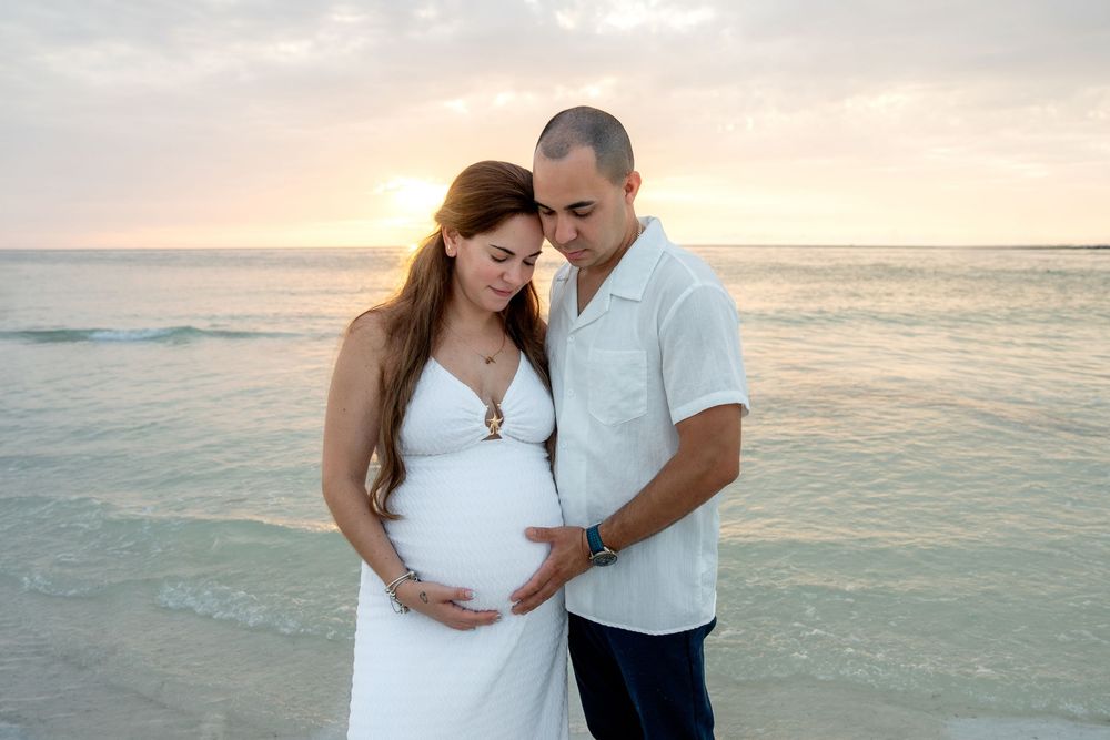 Pregnant woman in white dress and partner cradling baby bump on beach at sunset.