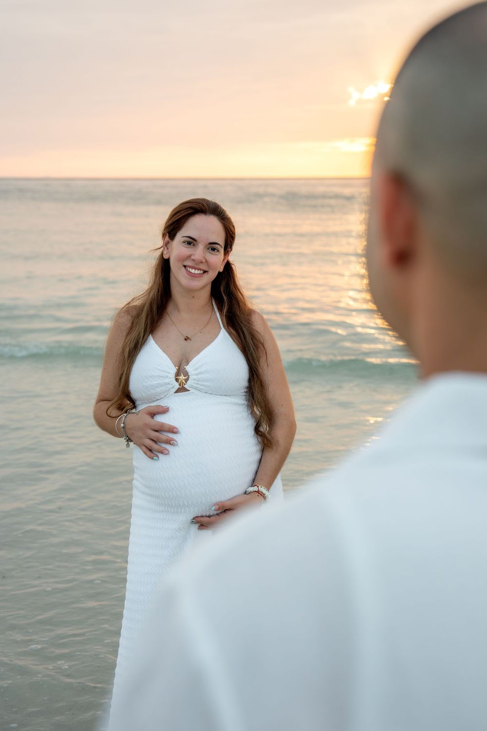 Smiling pregnant woman in white dress holds belly on beach at sunset with partner.