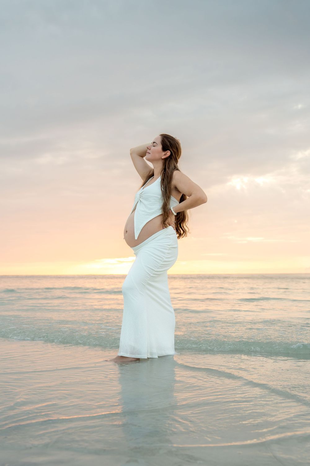 Pregnant woman in white dress stands in ocean water at stunning pink and purple sunset.