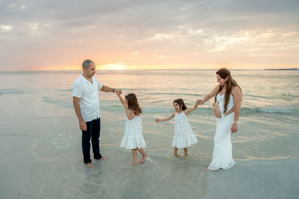 Family of four holds hands playing on beach in shallow water at colorful sunset.