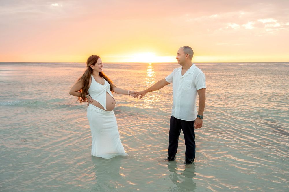 Expecting couple holds hands standing in ocean water facing each other at golden sunset.