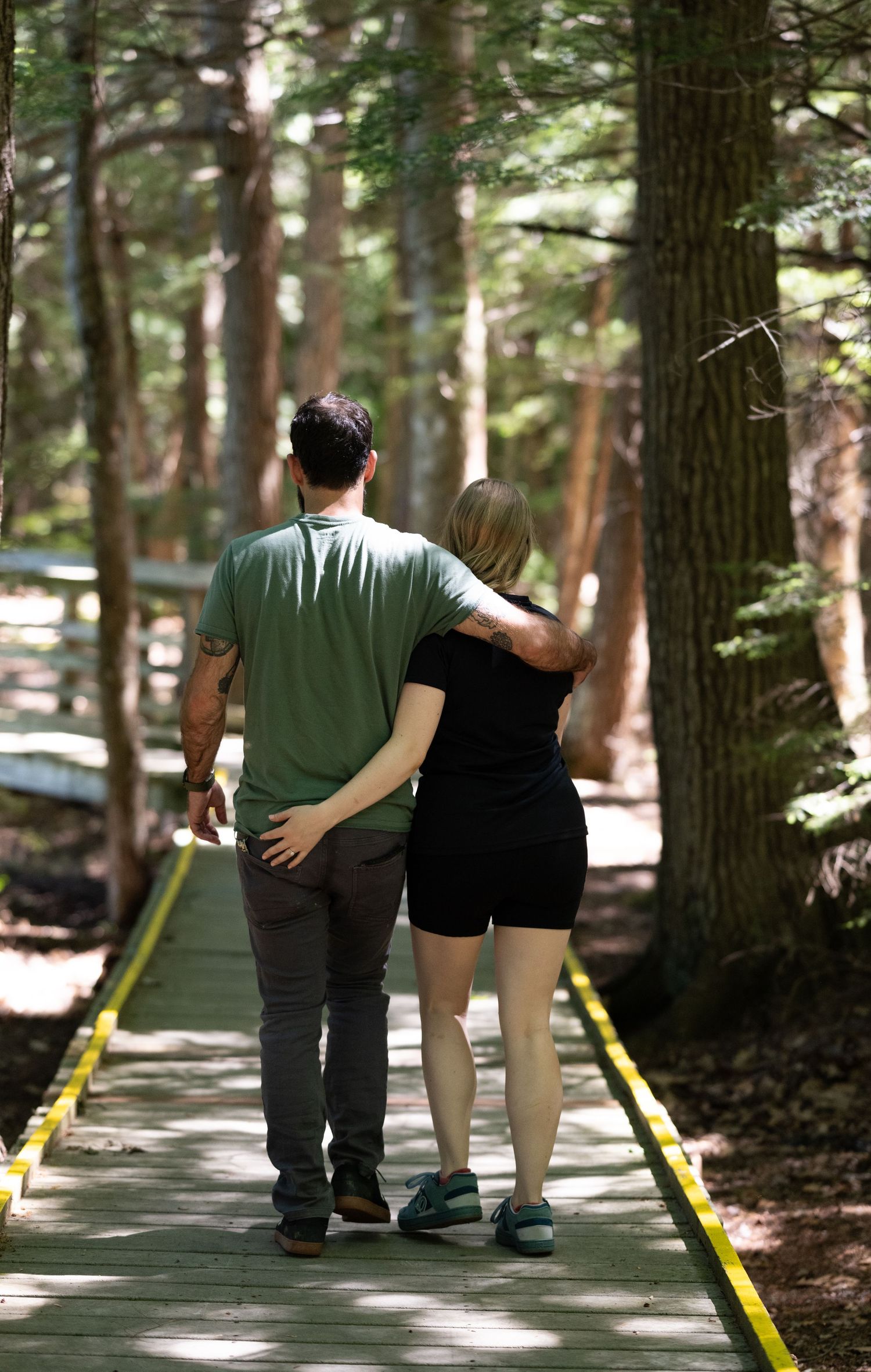Couple walking hand in hand along a wooden boardwalk path through a tall pine forest.