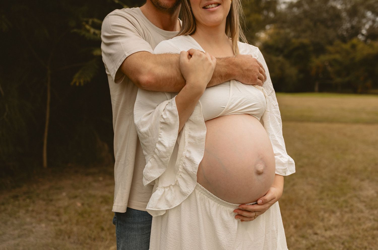 Outdoor maternity photoshoot featuring an expectant couple embracing in a sunny field at sunset.