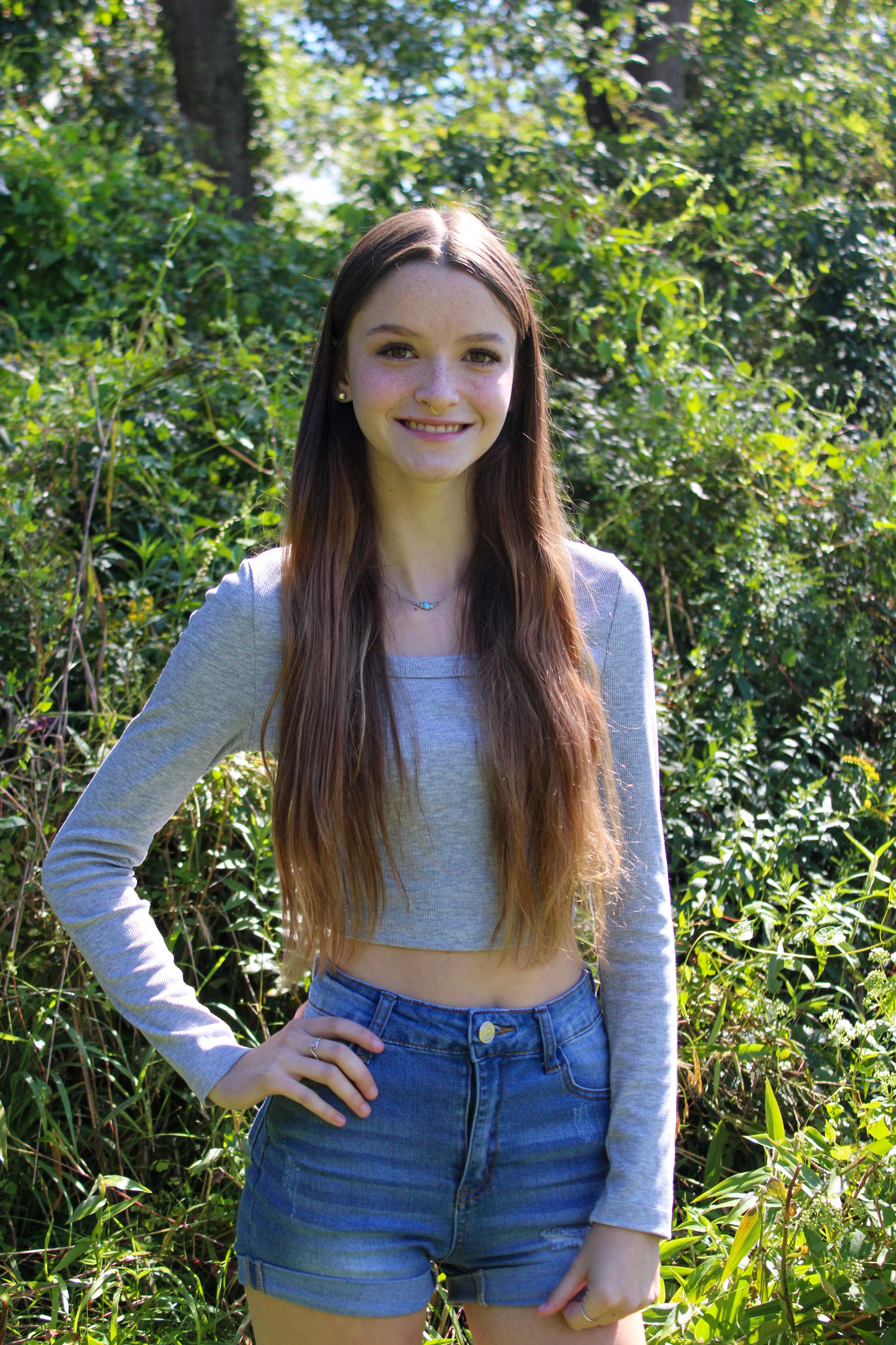 Person in casual outfit poses playfully in natural outdoor setting with green bushes.
