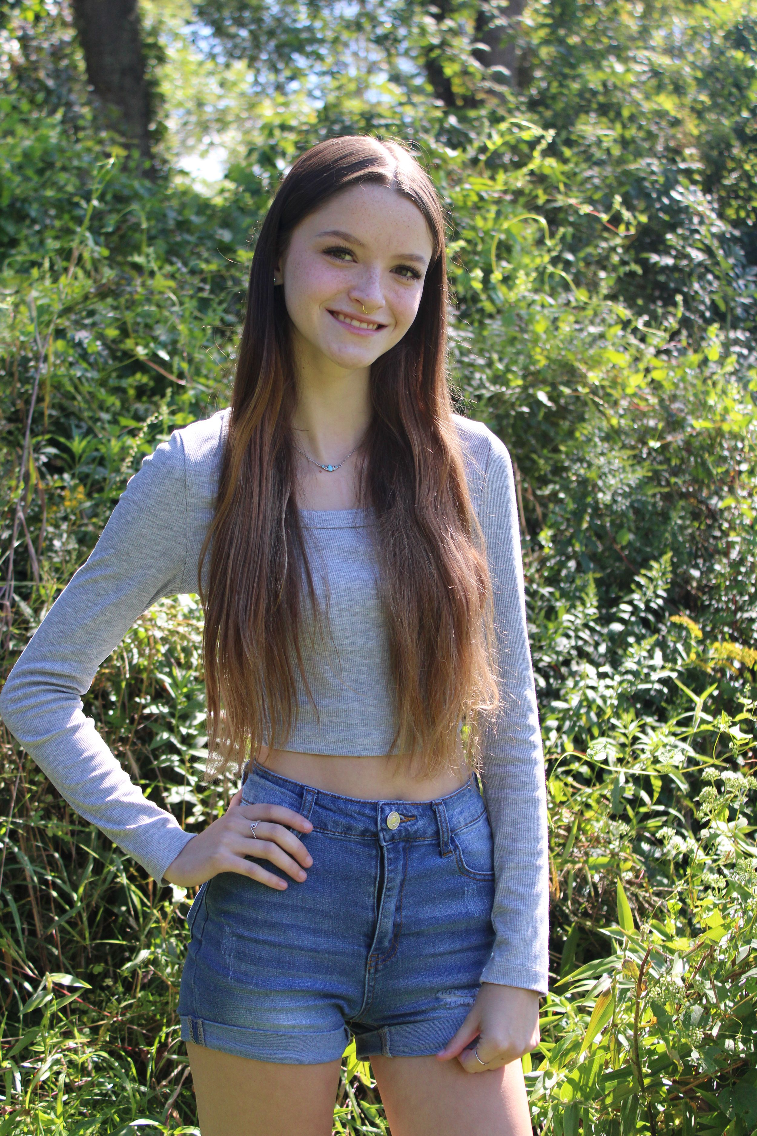 Person in grey crop top and denim shorts poses outdoors against green foliage background.