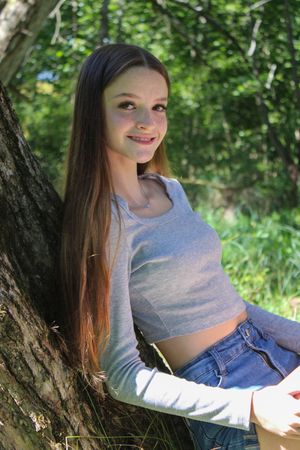 A person in a gray long-sleeve top and jeans poses casually near a tree in dappled sunlight.