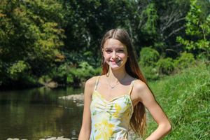 A person wearing a yellow floral romper poses by a rocky riverbank surrounded by lush green trees on a sunny day.
