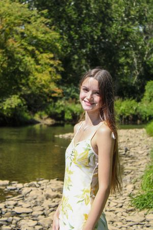 Someone poses in a white and yellow floral outfit by a river with a rocky shore and green foliage.