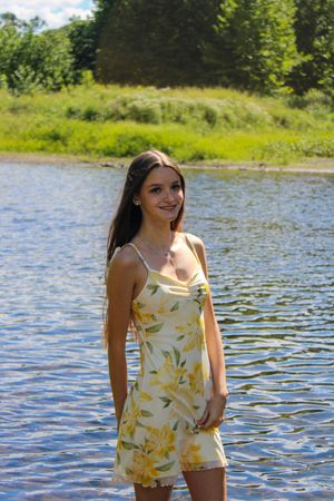 A bright summer day shows someone in a yellow floral romper standing in shallow water near a grassy shore.