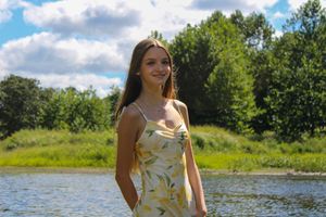 A summer landscape features someone in a floral outfit by a river with puffy white clouds overhead.