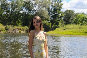 A bright summer scene shows someone in a yellow floral outfit by a river with green trees and blue skies.