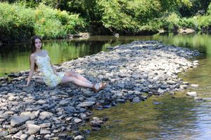 A woman in a summer dress relaxes on rocky riverbank surrounded by lush greenery.