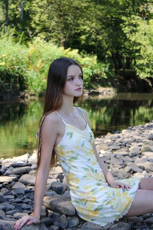 A woman in a white sundress with yellow flowers poses on rocky riverbank.