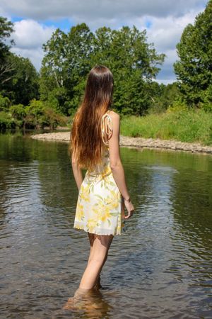 A woman in a yellow floral dress stands ankle-deep in a peaceful river.