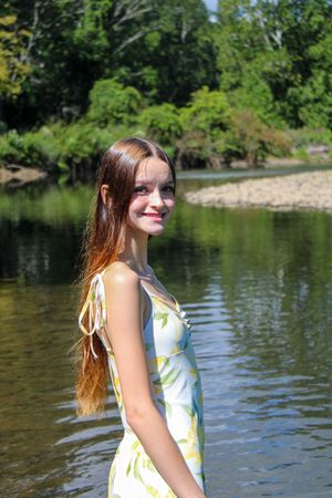 A woman in a summery dress poses by a tranquil river on a sunny afternoon.