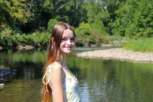 A woman in a white and yellow dress stands against a scenic river backdrop.
