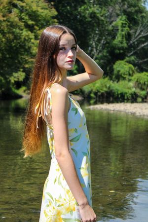 A woman in a floral sundress poses elegantly by a river on a bright summer day.
