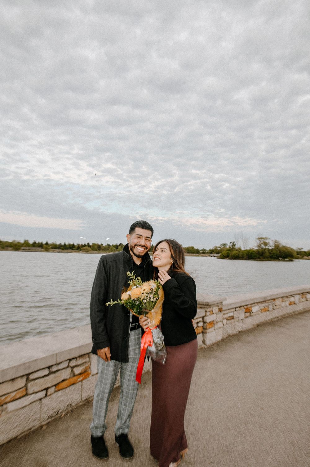 Couple sharing a romantic moment on a cloudy day at a waterfront pier with rustic railing and autumn flowers.