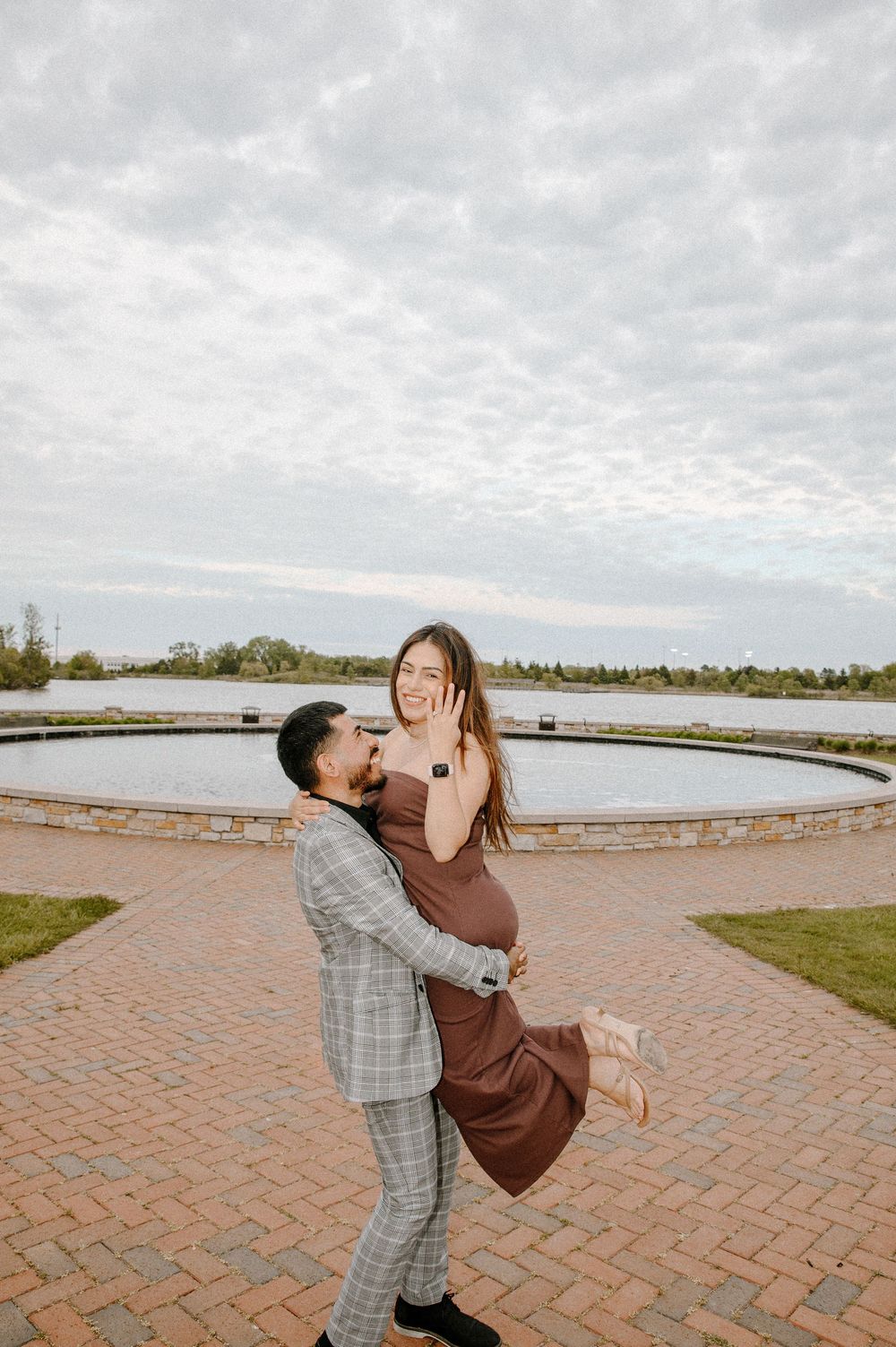 A couple in romantic embrace spins joyfully on brick pavement near a waterfront on an overcast day.