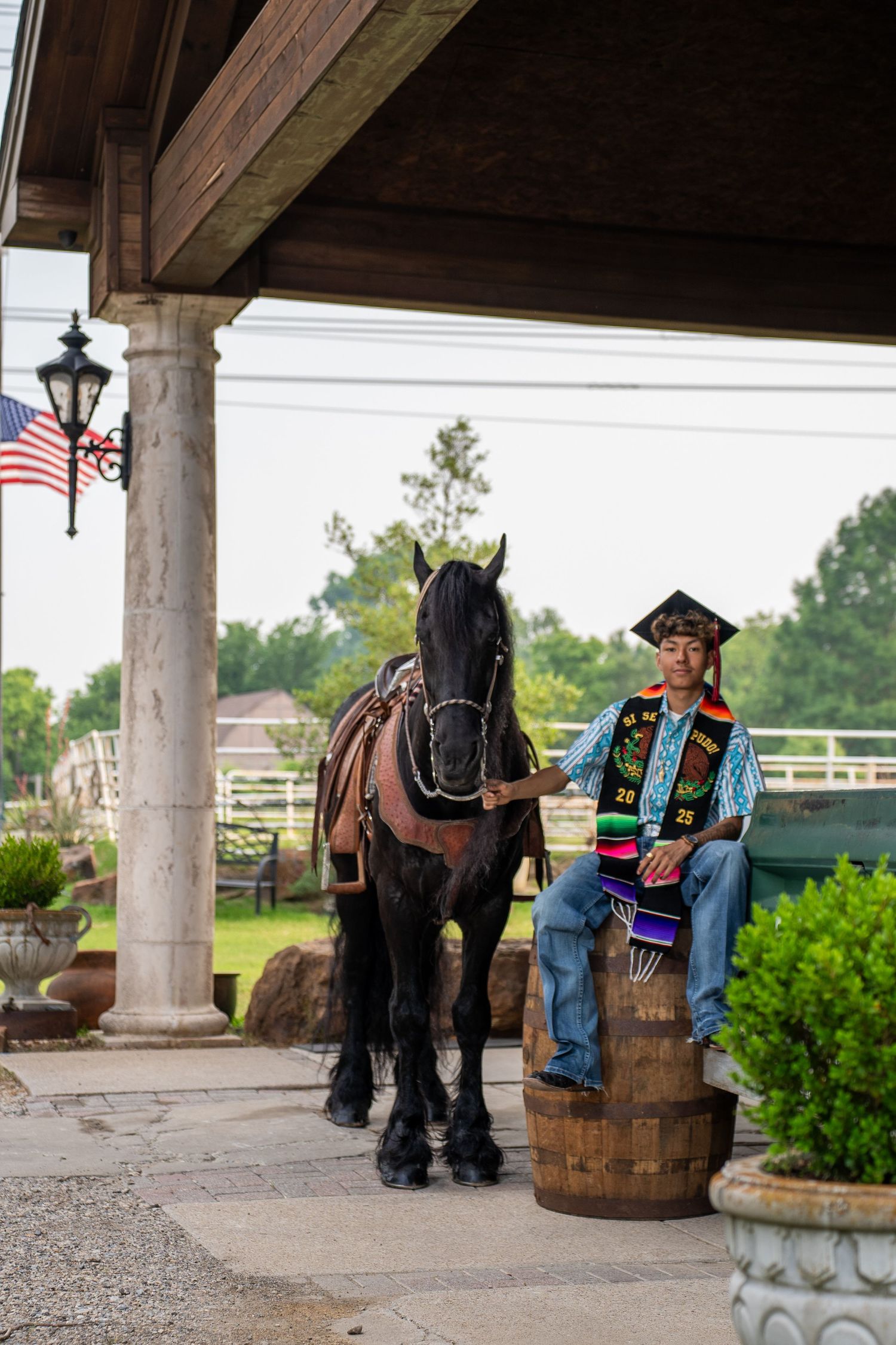 A graduate in academic regalia sits on a wooden barrel next to a black horse under a covered porch with an American flag.